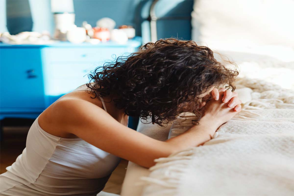 trauma bond photo • Dr. Toni Cooper Woman in a white tank top leans over a bed with her head bowed and hands clasped in front of her.