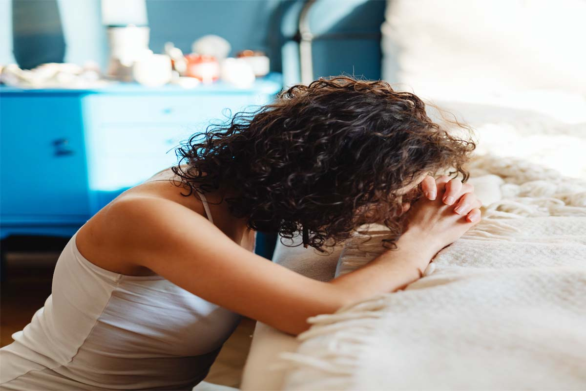 trauma bond photo Woman in a white tank top leans over a bed with her head bowed and hands clasped in front of her.